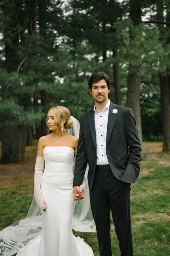 A bride in a strapless white gown and sheer gloves stands holding hands with a groom in a black suit and white shirt, both posing outdoors among tall green trees. by Justin Salem Meyer
