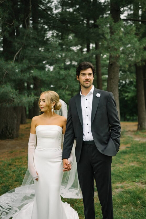A bride and groom stand hand in hand outdoors among tall trees. The bride wears a strapless white gown, sheer gloves, and a veil, while the groom wears a black suit with a white shirt and boutonnière. Both have a calm, serious expression. by Justin Salem Meyer