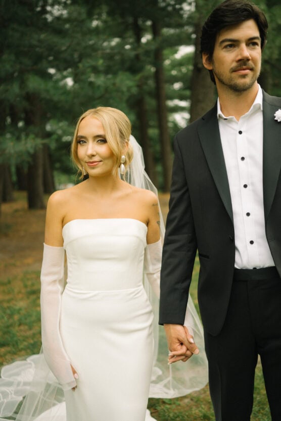 A bride in a white dress with sheer gloves and a veil holds hands with a groom in a black tuxedo. They stand outdoors in a wooded area, looking in different directions. by Justin Salem Meyer