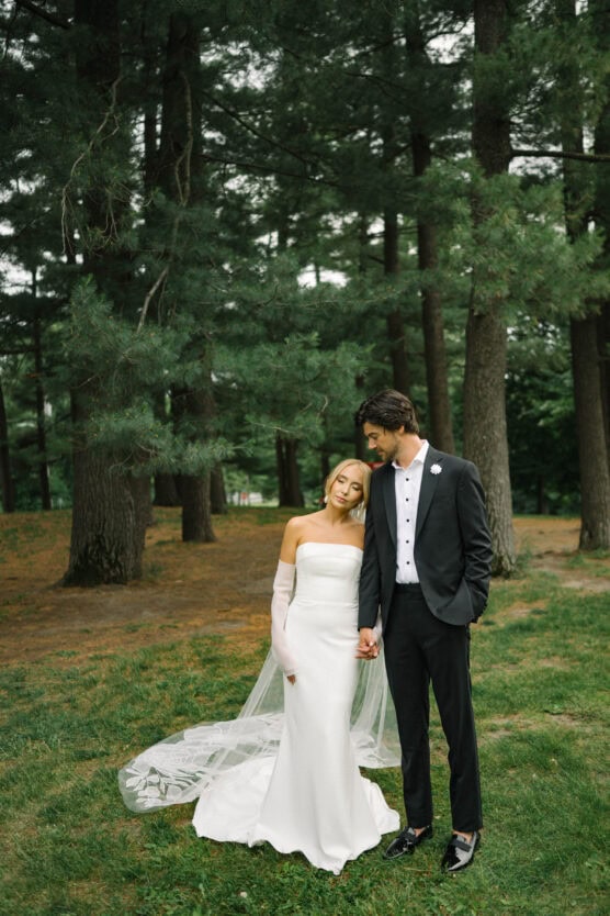 A bride in a strapless white gown and veil stands in a forest clearing, leaning her head on the groom’s shoulder. The groom, in a black suit, gently rests his head on hers as they hold hands, surrounded by tall green trees. by Justin Salem Meyer