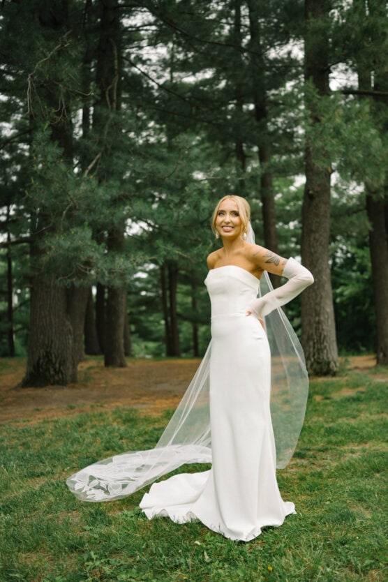 A woman in a strapless white wedding dress with sheer gloves and a veil stands smiling in a grassy area surrounded by tall pine trees. by Justin Salem Meyer