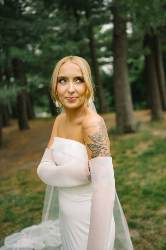 A bride in an off-the-shoulder white wedding dress poses outdoors among trees, with a floral tattoo visible on her upper arm and a sheer veil trailing behind her. She is smiling softly and looking to the side. by Justin Salem Meyer
