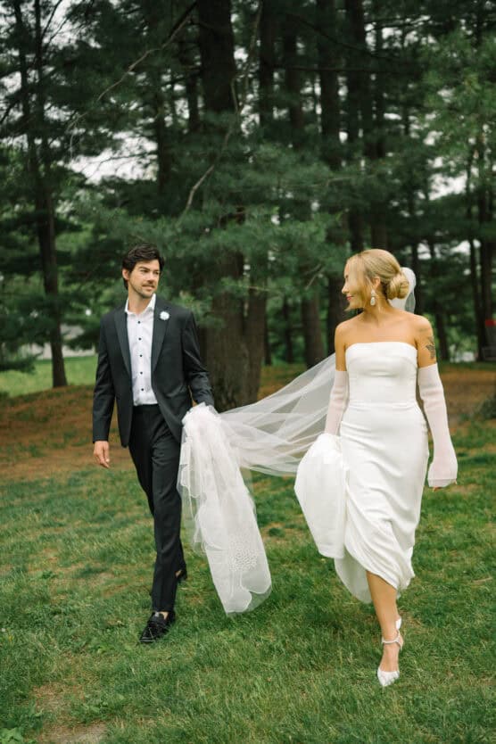A bride in a white dress and a groom in a black suit walk together on grass in a forested area. The groom holds part of the bride’s veil as they smile at each other. by Justin Salem Meyer