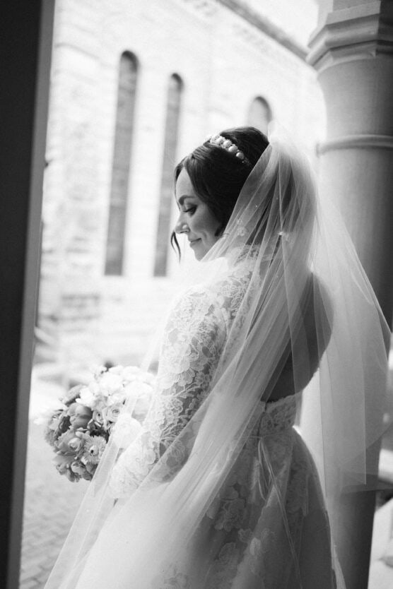 A bride in a lace gown and veil holds a bouquet and smiles, looking down. She stands near a stone building with arched windows, captured in soft black and white lighting. by Justin Salem Meyer