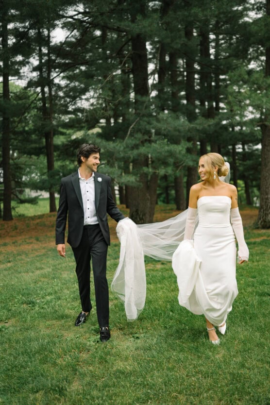 A bride in a white dress and gloves walks on grass, holding her gown as a groom in a black tuxedo holds her veil. They look at each other and smile, surrounded by tall pine trees. by Justin Salem Meyer