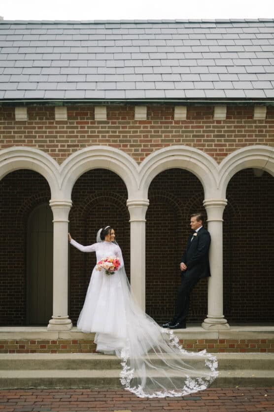 A bride in a long white gown and veil stands under an arched brick walkway, holding a bouquet of flowers, while a groom in a black suit leans casually against a column, gazing at her. by Justin Salem Meyer
