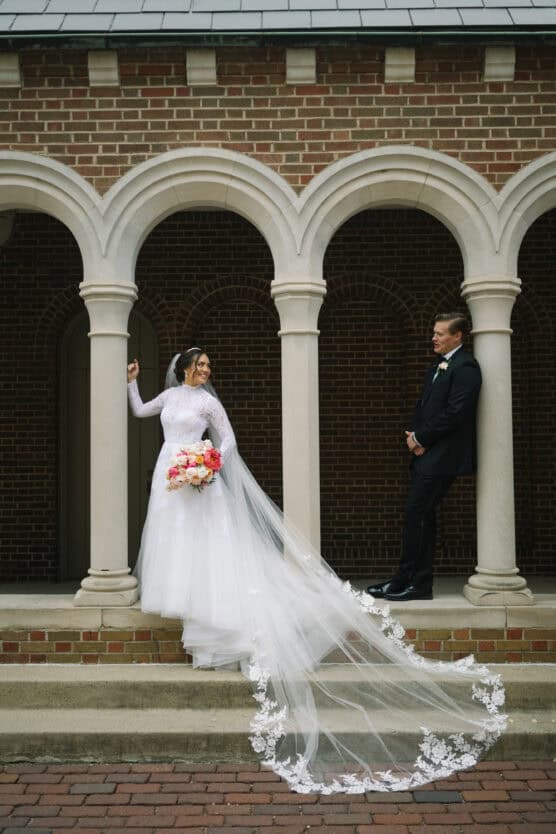 A bride in a long white gown with a lace train and holding a bouquet stands smiling under stone arches, while a groom in a black suit leans casually against a column, looking at her. by Justin Salem Meyer