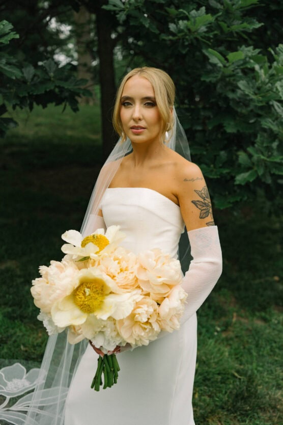 A bride in a strapless white gown and veil holds a large bouquet of pale yellow flowers. She stands outdoors in front of green foliage, showing a floral tattoo on her upper right arm. by Justin Salem Meyer