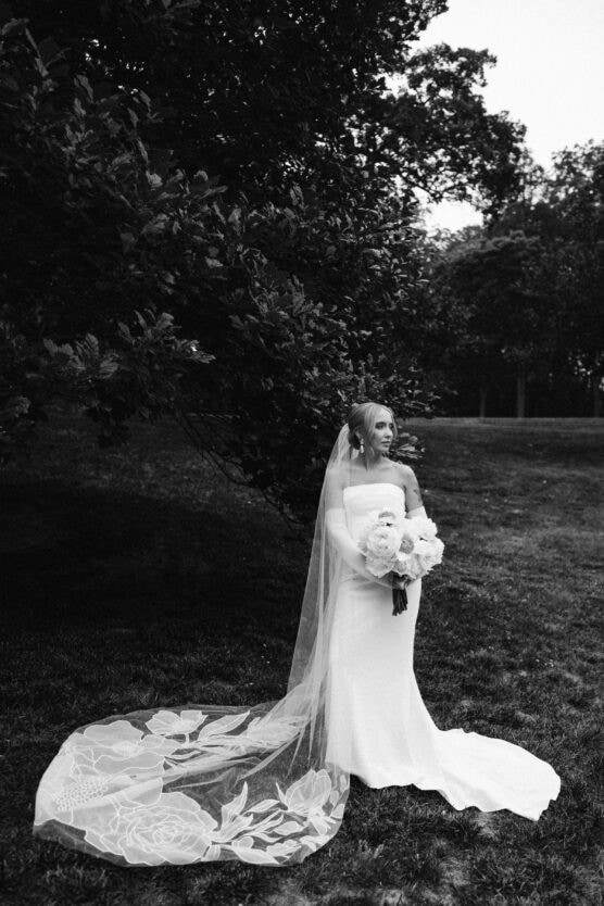 A bride in a strapless gown stands outdoors on grass, holding a bouquet of flowers. Her long veil with floral designs trails behind her. Trees and bushes fill the background. The photo is in black and white. by Justin Salem Meyer