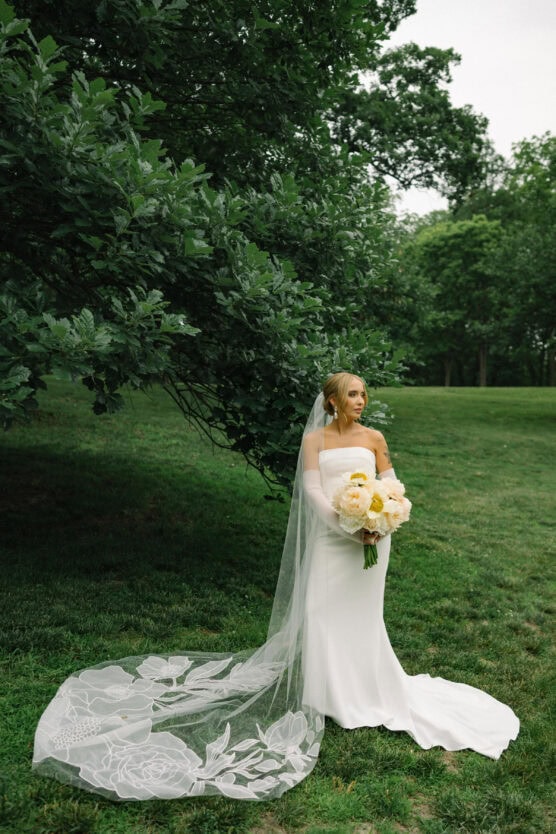 A bride in a strapless white gown and long veil with floral lace stands on grass holding a bouquet of yellow and white flowers, with lush green trees in the background. by Justin Salem Meyer