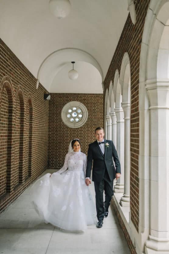 A bride in a white gown and veil walks hand in hand with a groom in a tuxedo down a brick corridor with arched windows and a round window in the background. by Justin Salem Meyer