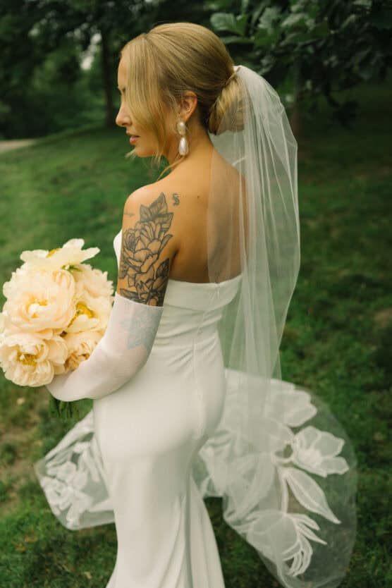 A bride in a strapless white wedding dress with a long veil stands outdoors, holding a bouquet of pale flowers. She has visible floral tattoos on her upper arm and is wearing large pearl earrings. by Justin Salem Meyer