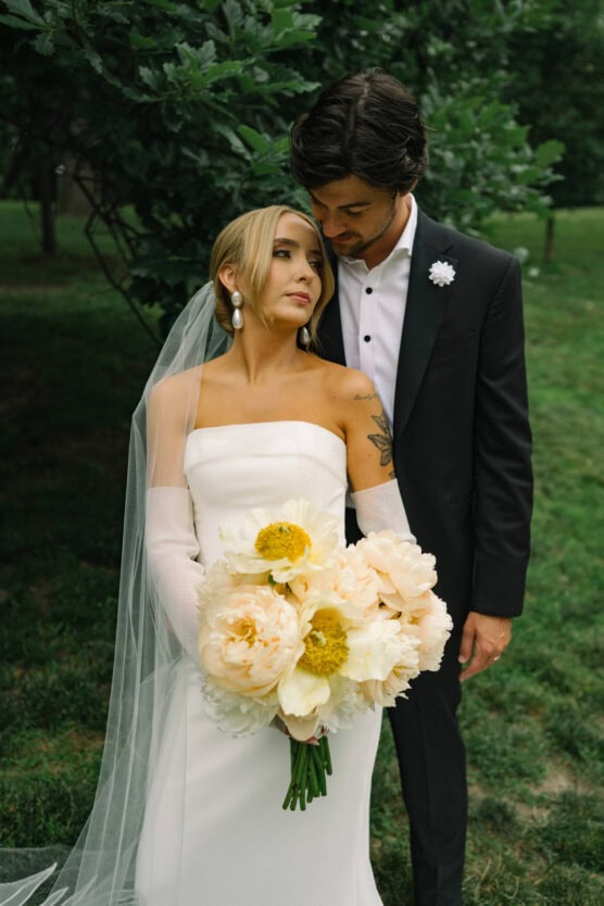 A bride in a white dress and veil holding a bouquet of pale yellow flowers stands closely with a groom in a black suit and white shirt. They look lovingly at each other outdoors amid greenery. by Justin Salem Meyer