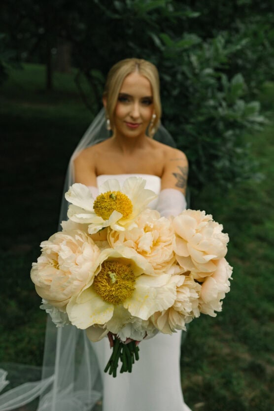 A bride in a white dress and veil holds a large bouquet of pale yellow peonies toward the camera, with greenery and blurred trees in the background. by Justin Salem Meyer
