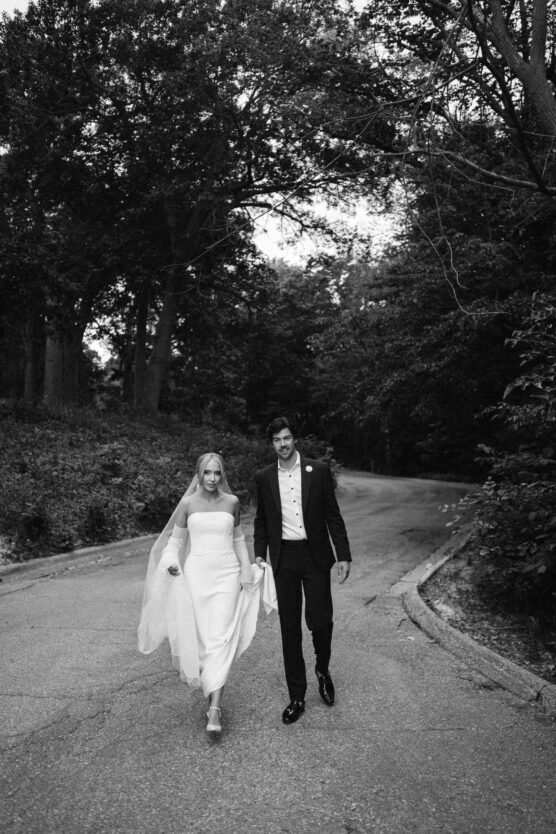 A bride in a strapless gown and veil walks hand in hand with a groom in a suit down a tree-lined road, both smiling. The photo is in black and white. by Justin Salem Meyer