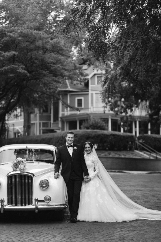 A bride and groom stand smiling beside a vintage car decorated with flowers. The bride wears a long gown and veil, while the groom is in a suit. Trees and a large, elegant house are visible in the background. by Justin Salem Meyer