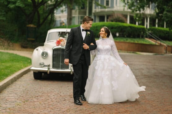 A bride and groom walk hand in hand on a cobblestone street. The bride wears a white, long-sleeved lace gown, and the groom wears a black tuxedo. A vintage white car decorated with flowers is parked nearby. by Justin Salem Meyer