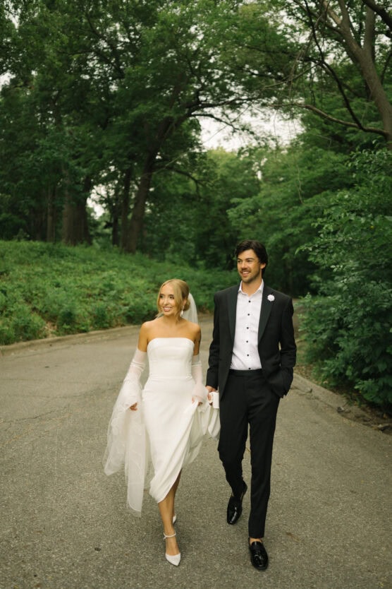 A bride in a white dress and a groom in a black suit walk hand in hand on a paved path surrounded by lush green trees, both smiling and looking happy. by Justin Salem Meyer