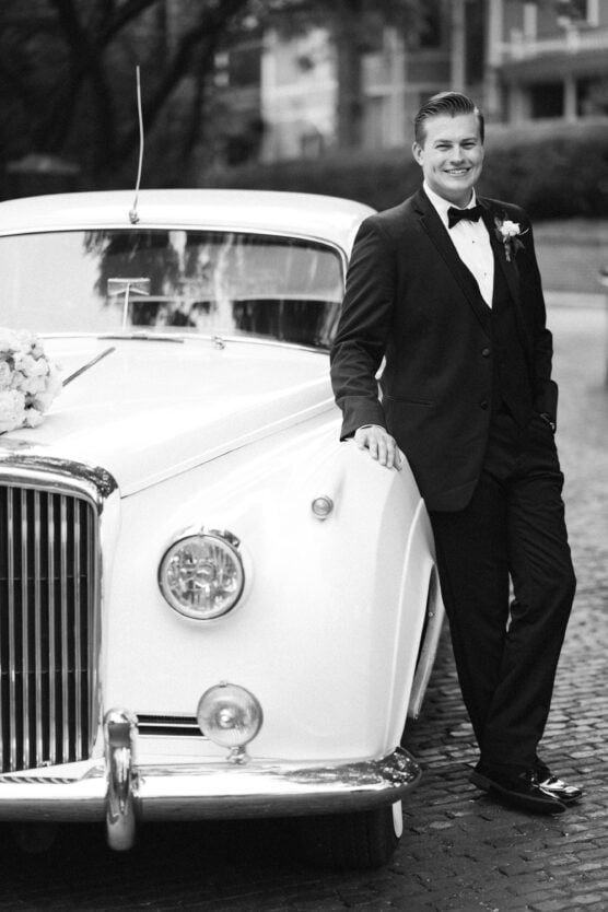 A man in a tuxedo smiles while leaning against a classic white car decorated with flowers. The scene appears formal, possibly at a wedding, with trees and buildings in the blurred background. The photo is in black and white. by Justin Salem Meyer