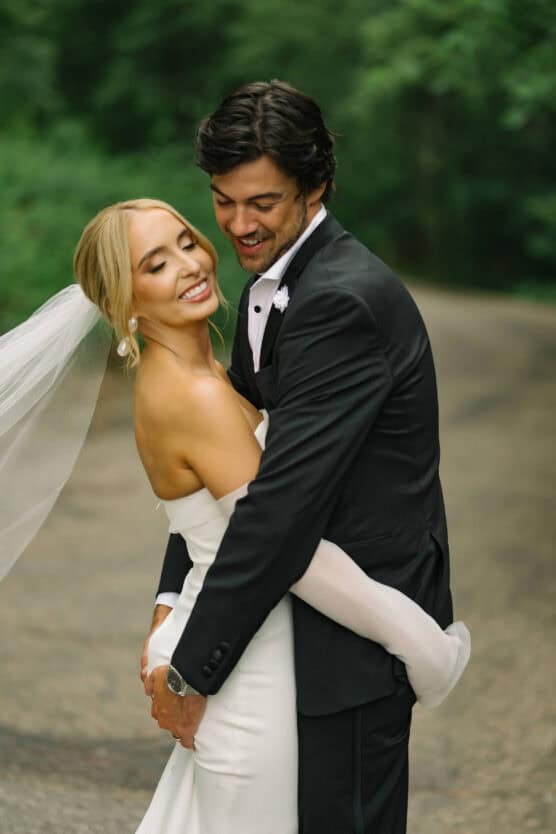 A bride in a white off-the-shoulder gown and veil smiles as she is embraced by a groom in a black tuxedo, both standing on a tree-lined outdoor path. by Justin Salem Meyer