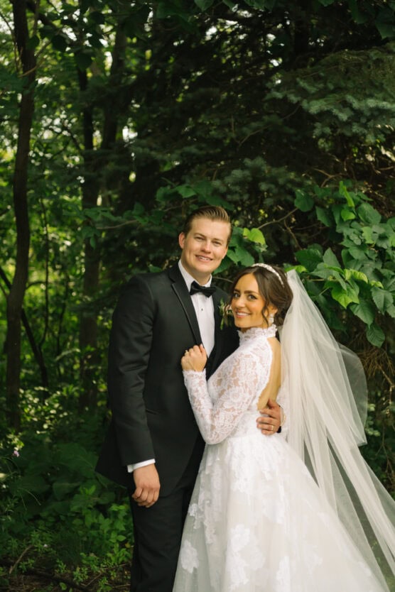 A bride and groom stand close together outdoors, smiling at the camera. The bride wears a long-sleeved lace wedding gown with a veil, and the groom wears a black tuxedo. Lush green foliage surrounds them. by Justin Salem Meyer