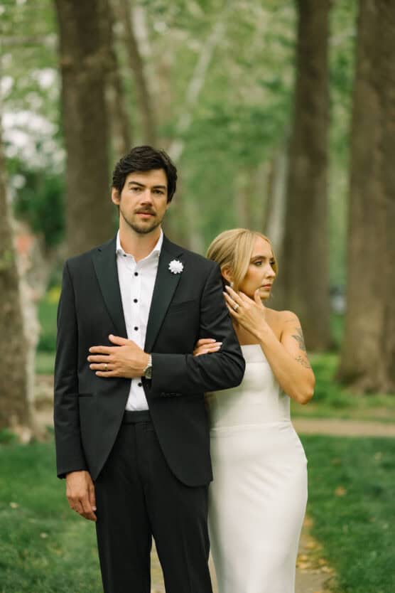 A bride in a strapless white dress stands beside a groom in a black suit, resting her hand on his shoulder. They are outdoors with tall trees and greenery in the background. by Justin Salem Meyer