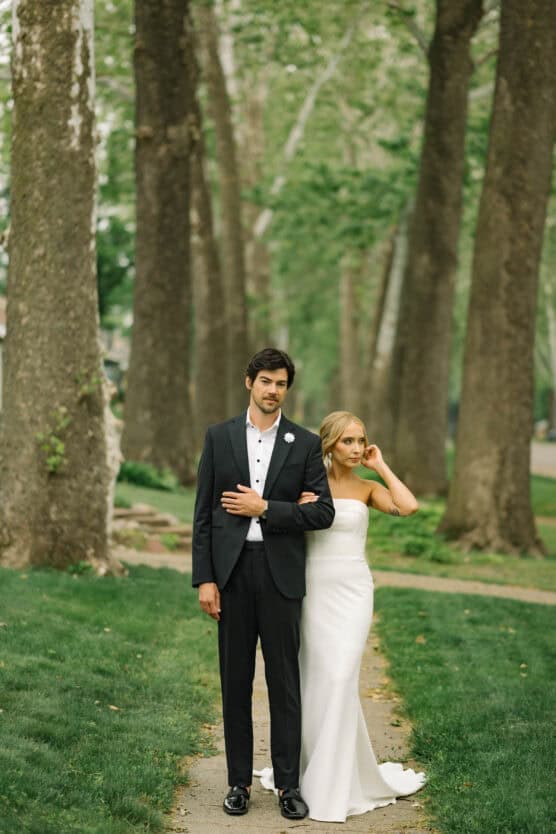 A bride in a strapless white gown stands arm-in-arm with a groom in a black suit and white shirt on a tree-lined path, surrounded by lush green grass and tall trees. by Justin Salem Meyer