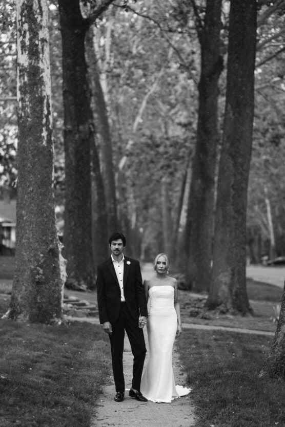 A couple dressed in formal wedding attire stands hand in hand on a tree-lined path. The groom wears a dark suit and the bride wears a strapless white gown. The image is in black and white. by Justin Salem Meyer