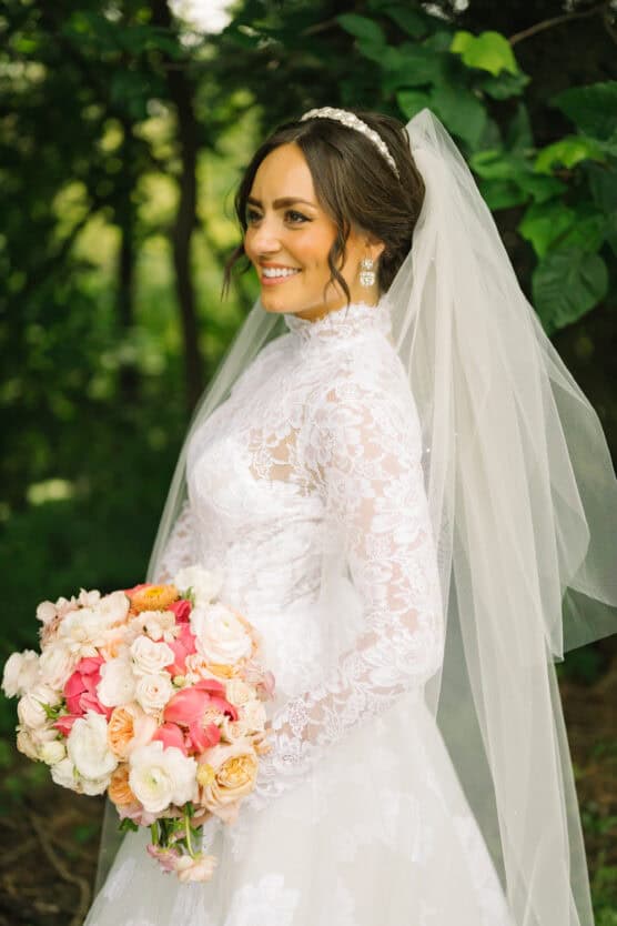 A bride in a long-sleeve lace wedding dress and veil stands outdoors, holding a bouquet of pink and white flowers, smiling and looking to the side. Lush greenery is visible in the background. by Justin Salem Meyer