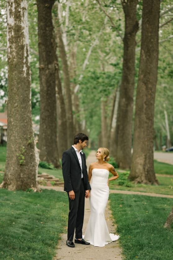 A bride in a strapless white dress and a groom in a black suit hold hands and look at each other while standing on a tree-lined path in a lush, green park. by Justin Salem Meyer