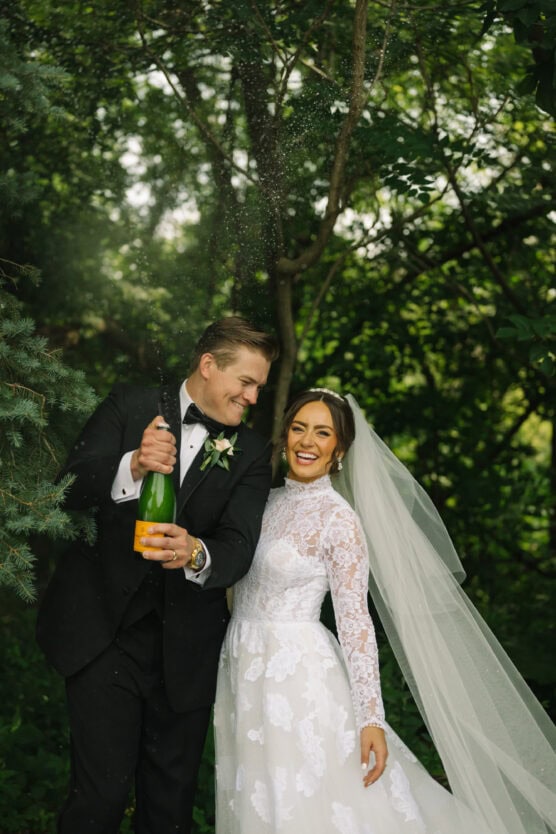 A groom in a black tuxedo pops a champagne bottle next to his smiling bride in a long lace wedding dress and veil, standing together outdoors amid lush green trees. by Justin Salem Meyer