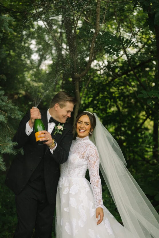 A groom in a black tuxedo pops a champagne bottle while standing next to his smiling bride in a white lace wedding dress and veil. They are outdoors surrounded by lush green trees. by Justin Salem Meyer