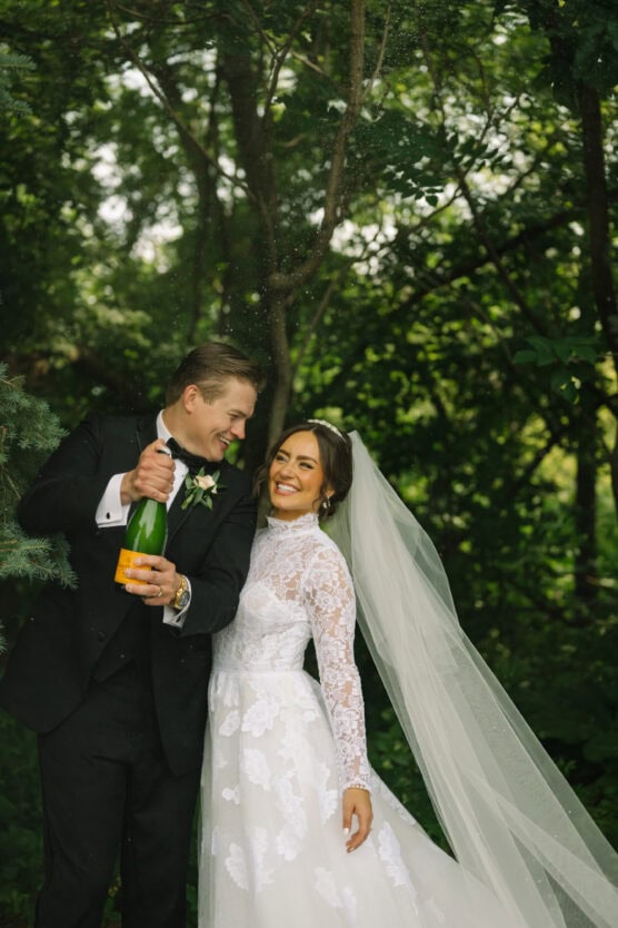 A groom in a black tuxedo pops a champagne bottle next to his smiling bride in a long-sleeved lace wedding dress and veil, both standing in a lush, green outdoor setting. by Justin Salem Meyer