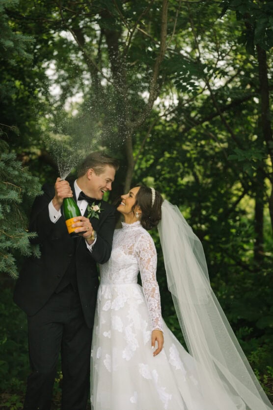 A newlywed couple stands outdoors in a lush, green forest. The groom, wearing a black suit, pops a bottle of champagne, while the bride, in a white lace gown and veil, smiles up at him. Champagne sprays in the air as they celebrate. by Justin Salem Meyer
