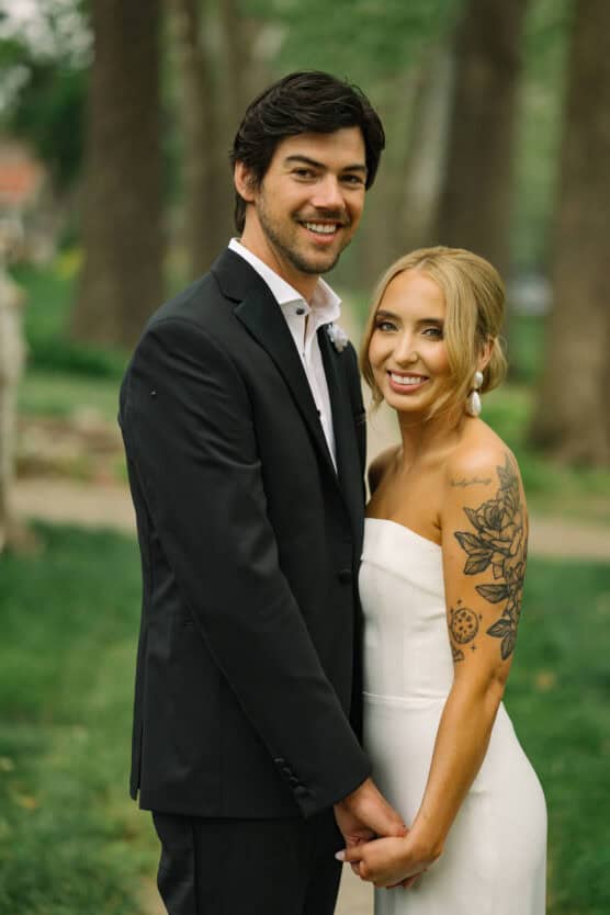 A smiling bride and groom stand outdoors holding hands. The groom wears a black suit, while the bride wears a strapless white dress and has a floral tattoo on her arm. They are surrounded by greenery and trees. by Justin Salem Meyer