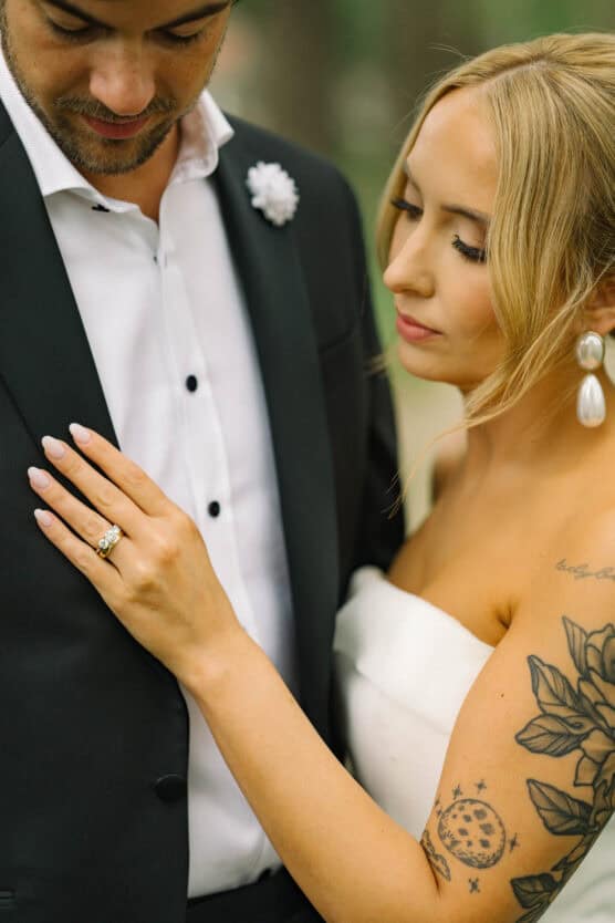 A bride with blonde hair and visible tattoos gently touches the chest of a groom in a black suit and white shirt. She wears pearl earrings and a ring, both appear calm and close, suggesting an intimate wedding moment. by Justin Salem Meyer