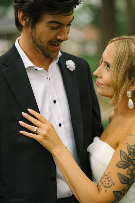 A bride with tattoos and pearl earrings lovingly gazes at a groom in a black suit. She rests her hand with a wedding ring on his chest; both appear happy and close, standing outdoors. by Justin Salem Meyer