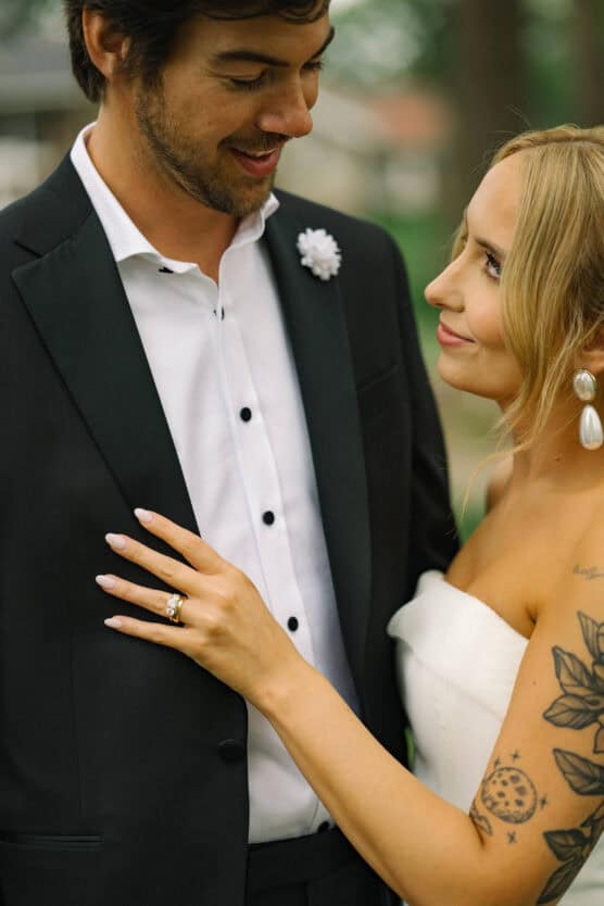 A bride with long blonde hair and floral tattoos on her arm lovingly holds her groom’s jacket. The groom wears a black suit and white shirt. They smile and gaze at each other outdoors, appearing happy and in love. by Justin Salem Meyer