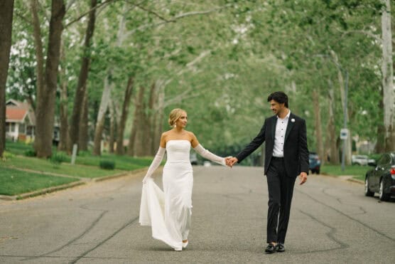 A bride in a white gown and a groom in a black tuxedo hold hands and walk down a tree-lined street, smiling at each other. The scene is peaceful, with green trees and houses in the background. by Justin Salem Meyer