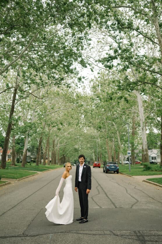 A bride in a white dress and a groom in a black tuxedo stand together in the middle of a tree-lined residential street, smiling on a calm, overcast day. by Justin Salem Meyer
