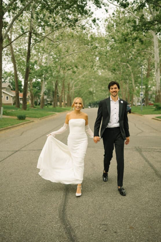 A bride in a white gown and a groom in a black tuxedo walk hand in hand down a tree-lined street, smiling at the camera. by Justin Salem Meyer