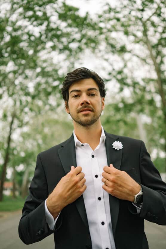 A man in a black suit and white shirt stands outdoors, buttoning his jacket. He wears a white flower on his lapel and looks confidently at the camera, with green trees blurred in the background. by Justin Salem Meyer
