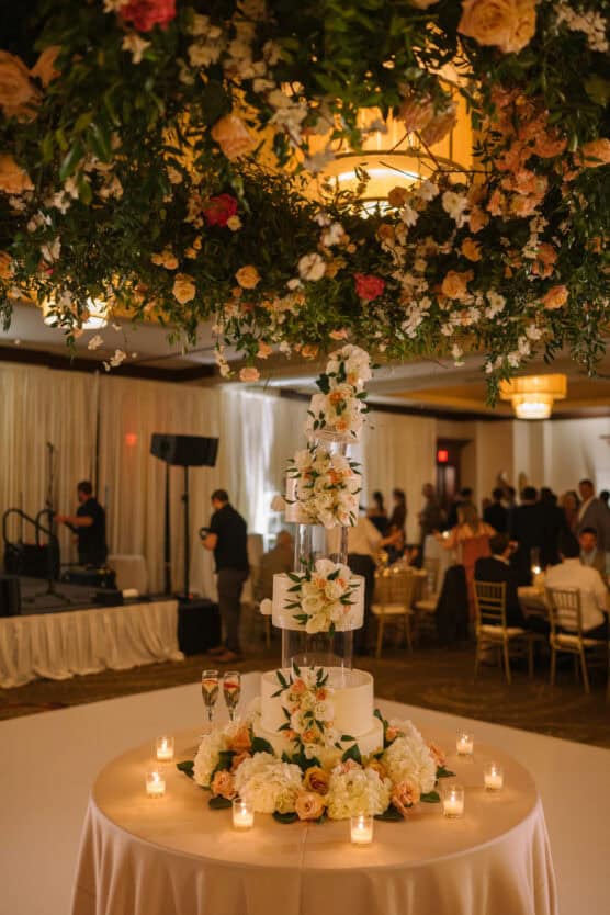 A multi-tiered white wedding cake decorated with flowers sits on a round table with candles, under a hanging floral arrangement. People are seated and mingling in a softly lit reception hall. by Justin Salem Meyer