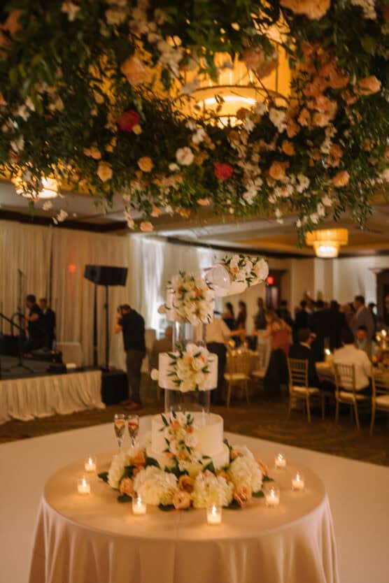 A three-tiered wedding cake decorated with white and blush flowers sits on a round table surrounded by candles; floral arrangements hang above, with guests and tables visible in the softly lit background. by Justin Salem Meyer