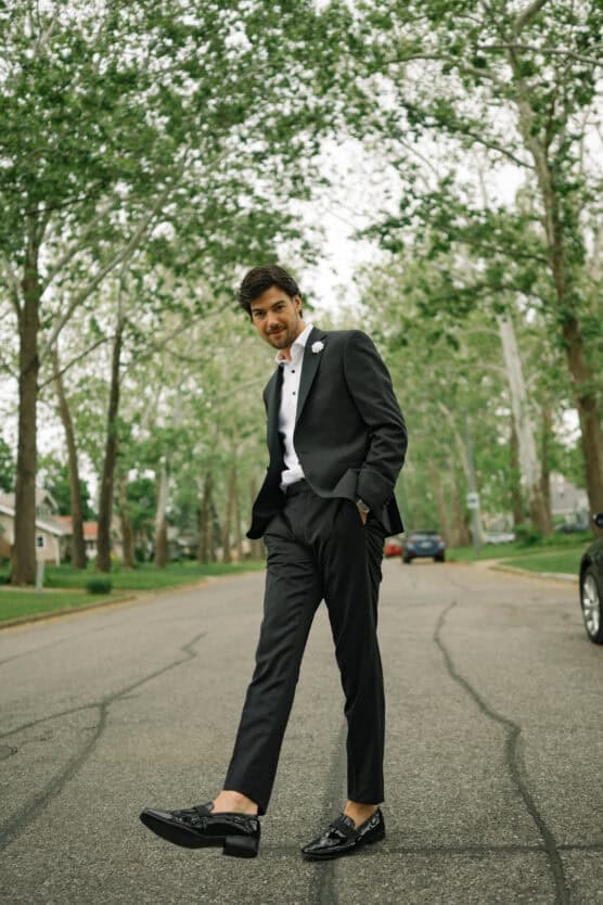 A man in a black suit and dress shoes stands confidently in the middle of a tree-lined suburban street, hands in pockets, with a relaxed expression and slightly forward stride. by Justin Salem Meyer
