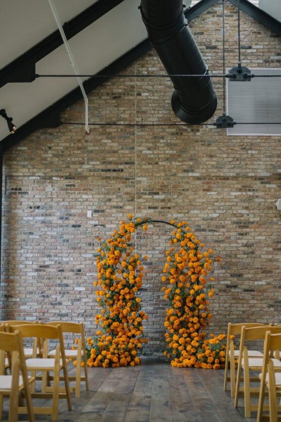 A modern indoor wedding setup with wooden chairs facing an arch decorated with vibrant orange marigold flowers, set against an exposed brick wall and vaulted ceiling. by Justin Salem Meyer