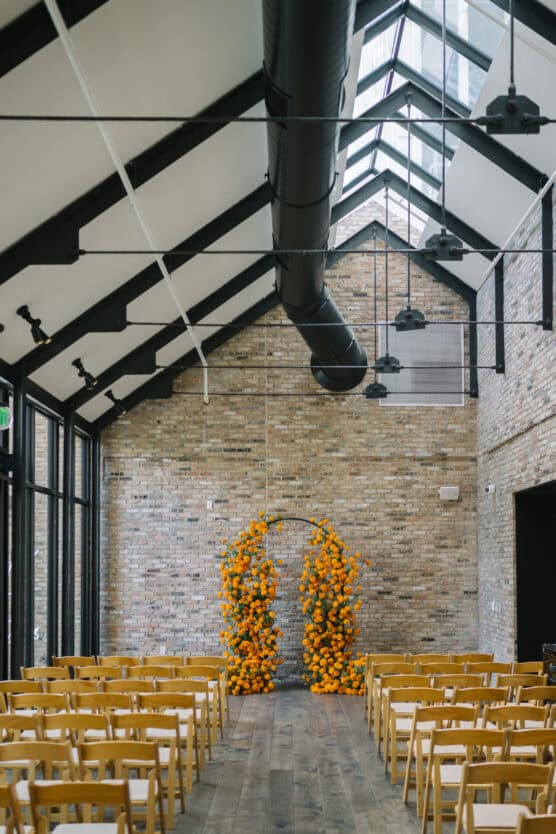 A bright, modern indoor wedding venue with rows of wooden chairs facing a floral arch covered in vibrant orange flowers, set against a tall brick wall and large windows with a high, pitched ceiling. by Justin Salem Meyer