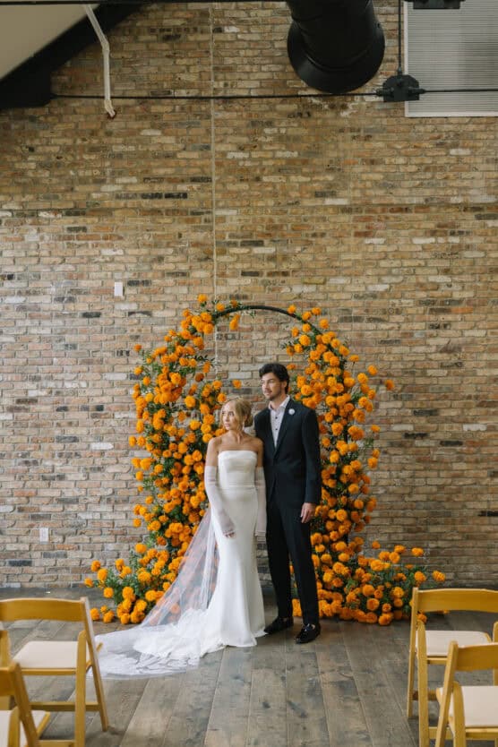 A bride and groom stand together in front of a circular floral arch made of vibrant orange flowers, set against a rustic brick wall. Yellow chairs are arranged in rows on a wooden floor. by Justin Salem Meyer