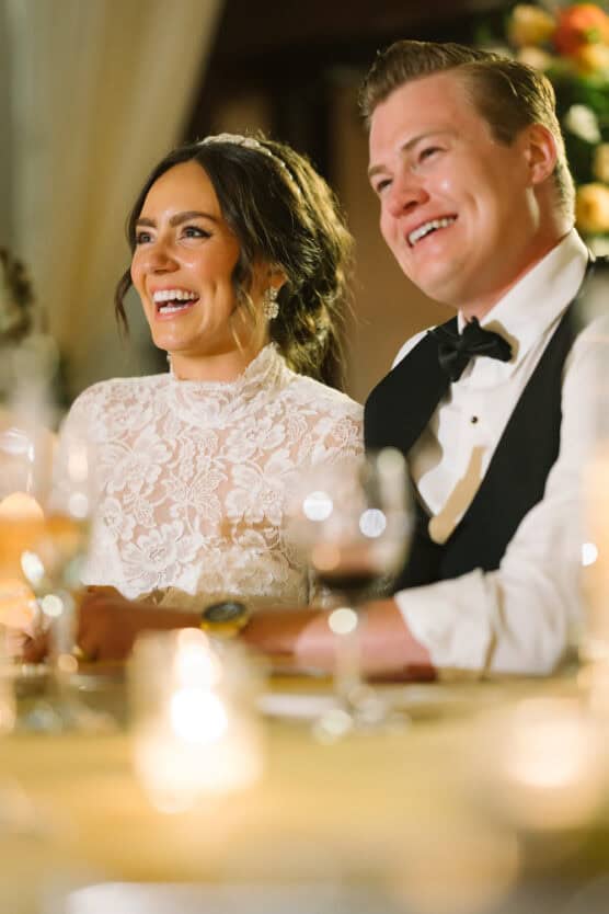 A bride in a lace dress and a groom in a tuxedo sit side by side, smiling and laughing at a warmly lit wedding reception with candles and blurred floral decorations in the background. by Justin Salem Meyer