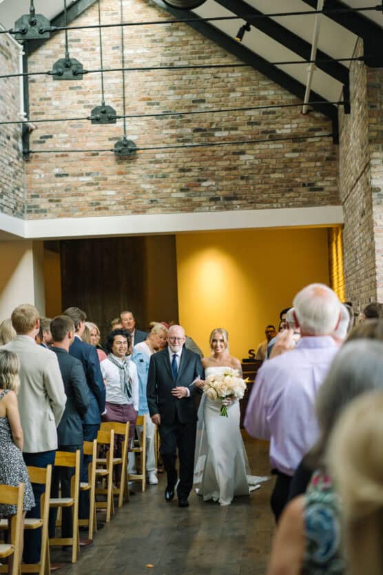 A bride in a white dress holding a bouquet walks down the aisle with an older man in a suit, surrounded by seated and standing wedding guests in a venue with exposed brick walls and high ceilings. by Justin Salem Meyer